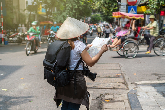 A person wearing a conical straw hat and carrying a backpack stands on a sidewalk in a busy Southeast Asian street.