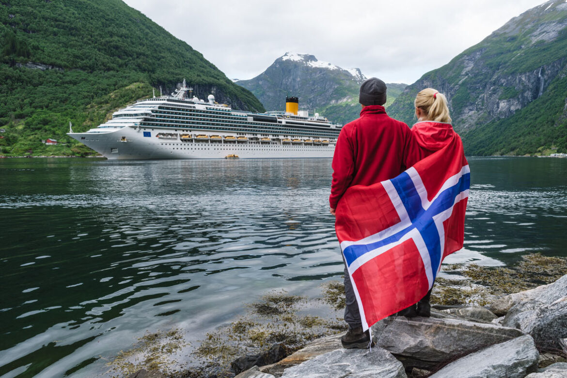 Couple of travelers with a Norwegian flag look at the Cruise liner. Fjord Geirangerfjord near Geiranger city.